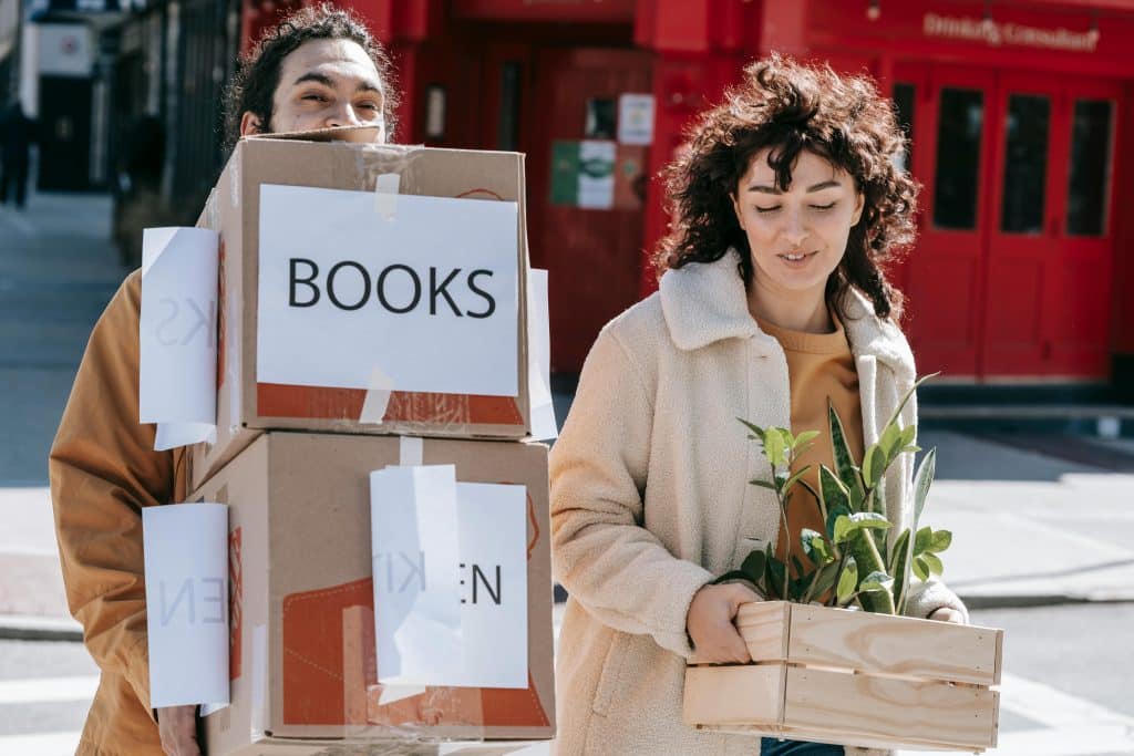 Man holding multiple moving boxes walking behind a woman carrying a plant