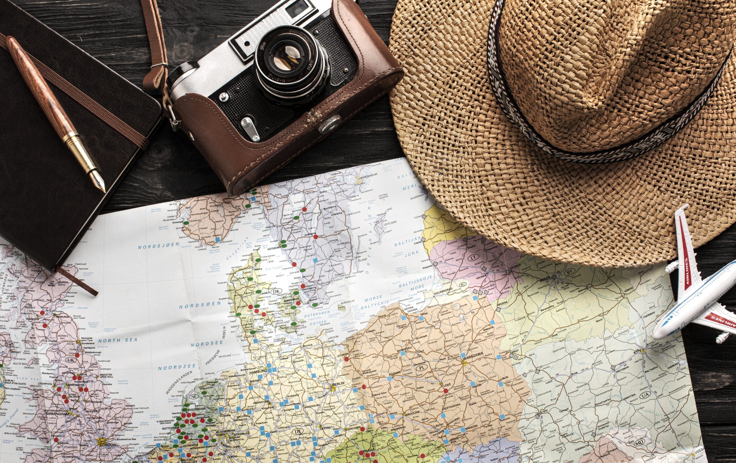 Map, model airplane, camera, black notebook with a brown fountain pen laying on top and a straw hat on top of a desk