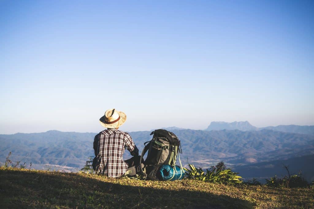 Man with a backpack sitting on top of a mountain with his back to the camera and looking down at a city